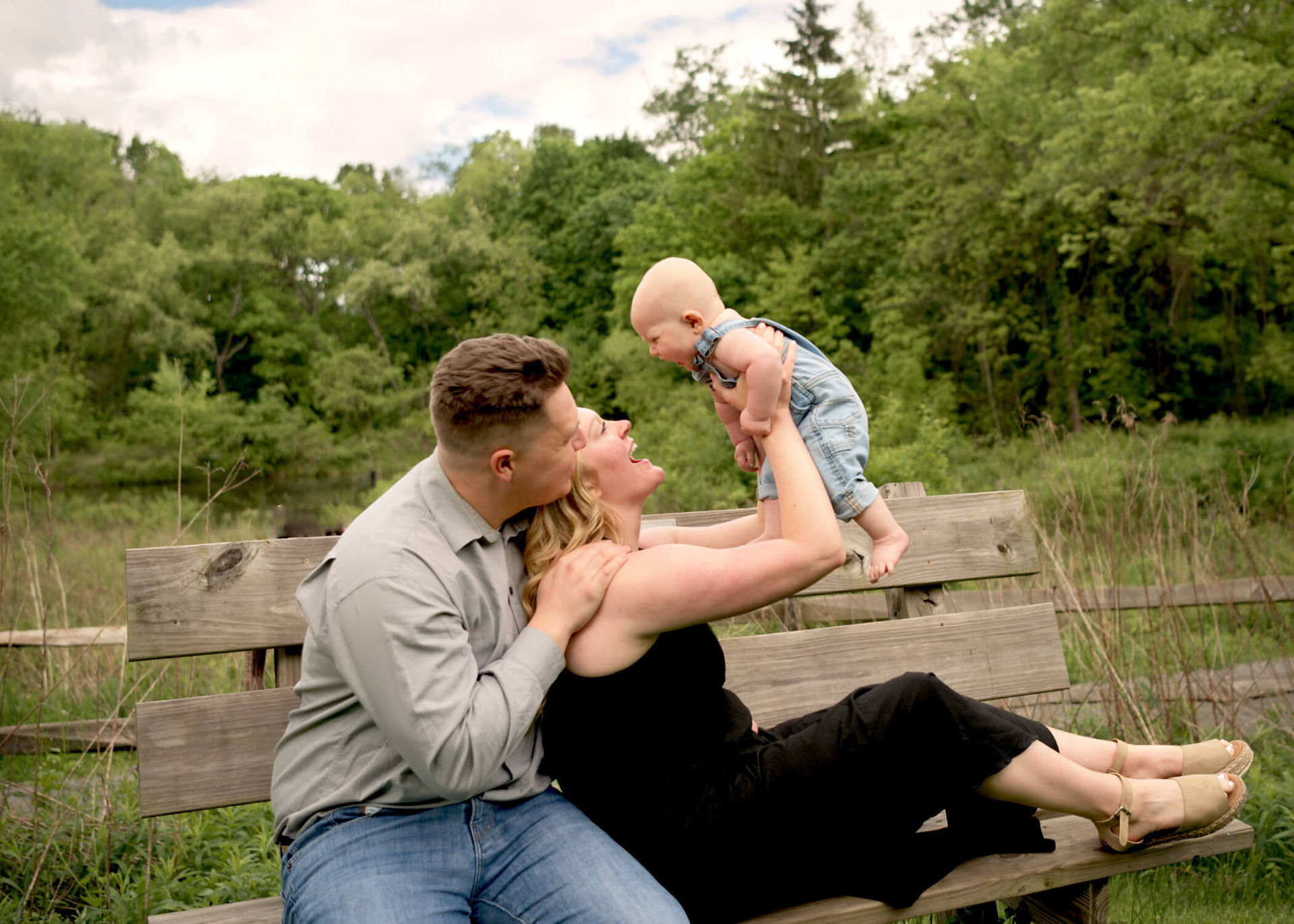A mother holds her baby son above her head while sitting on a bench and her husband sitting beside her. 
