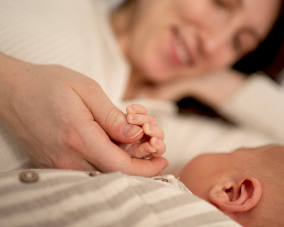 A mother is laying down with her newborn son on a bed and holding his tiny fingers while smiling.