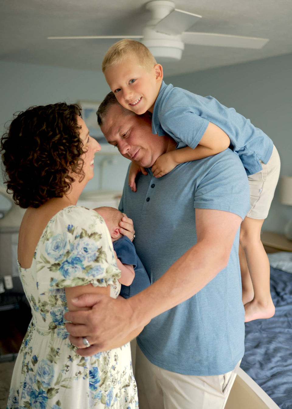 A family is playfully interacting in the master bedroom with a newborn and the older brother on the dad's back.