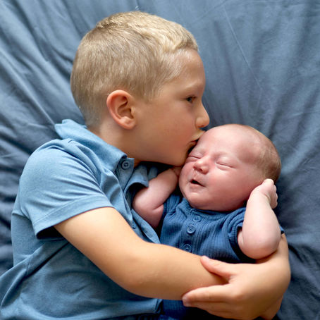 A young boy is holding and kissing the forehead of his newborn baby brother while laying on a bed. 