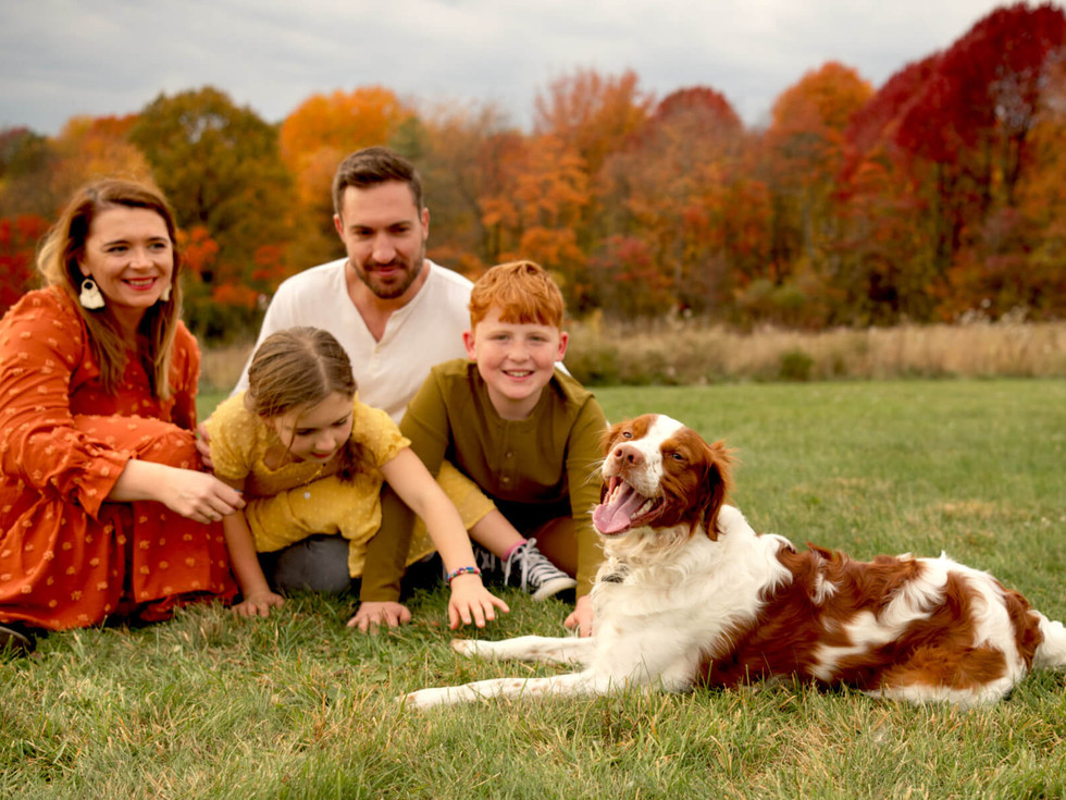 A family of four sits in a field with their dog with vibrant fall colors around them.