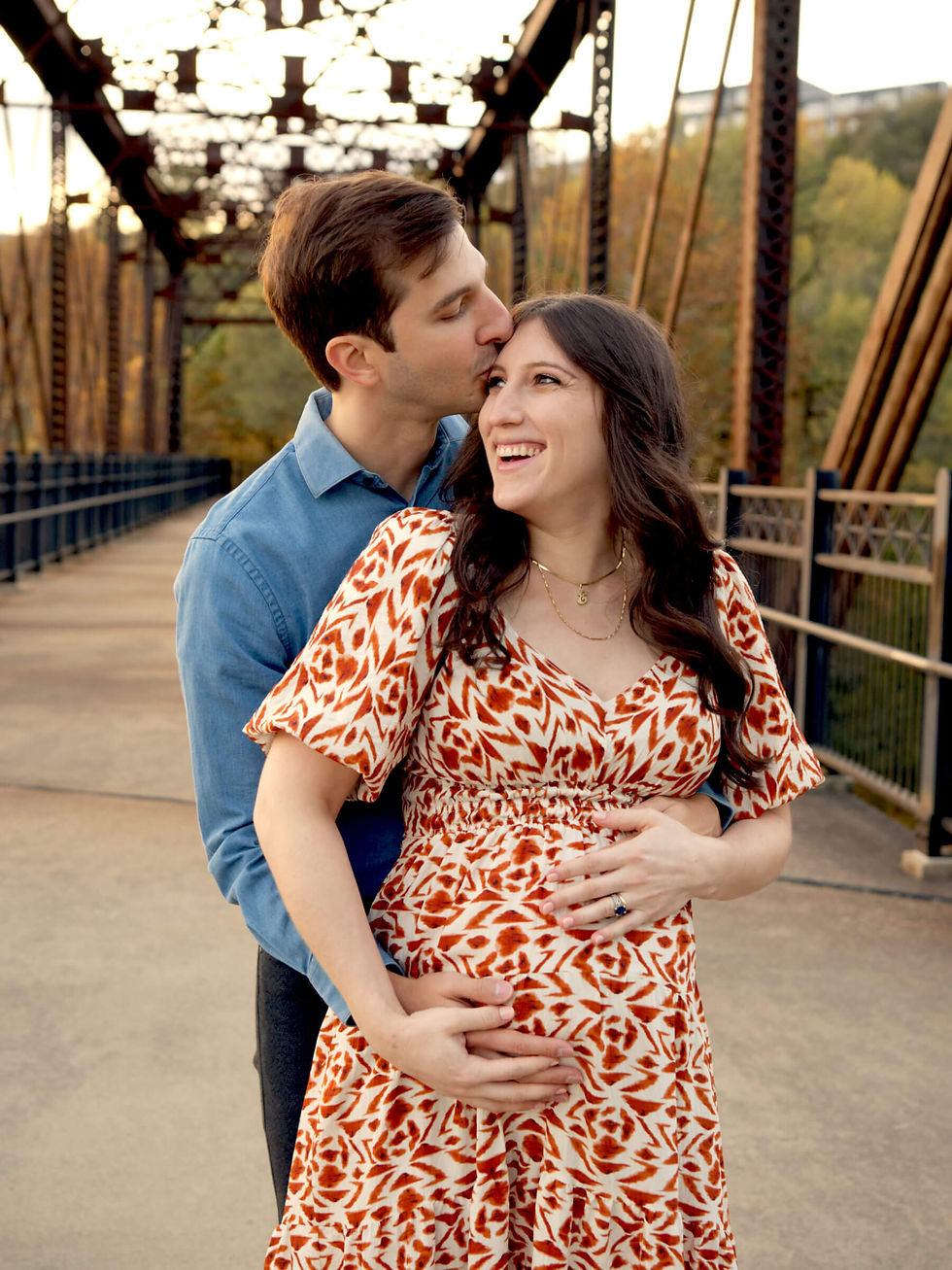A pregnant mother and her husband hug and laugh while standing on a bridge in Pittsburgh, PA.
