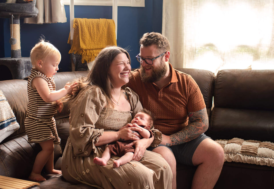 A mother laughs as her toddler plays with her hair on the couch and she holds a baby boy, sitting next to her husband.