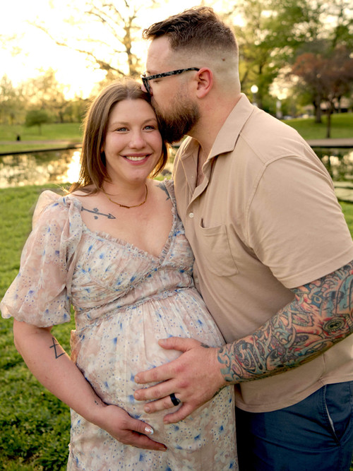 A pregnant mother stands with her husband, his hand on her baby bump, while standing in Allegheny Commons Park.