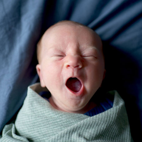 A newborn baby boy is widely yawning while laying on a bed.
