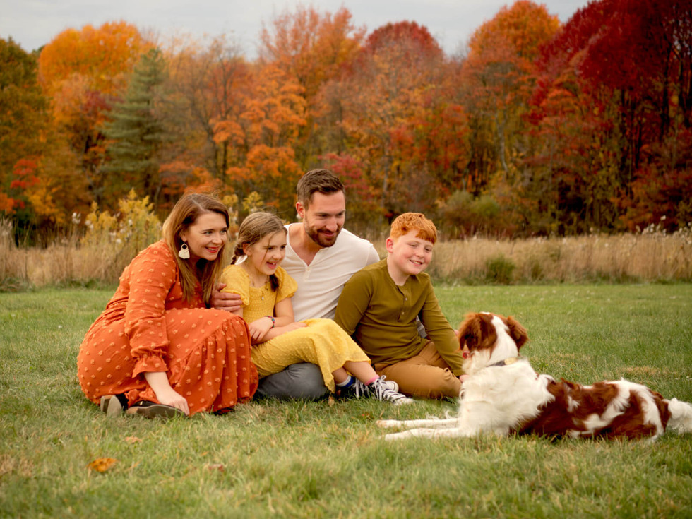 A family of four happily smiles at their dog while sitting in a field. 