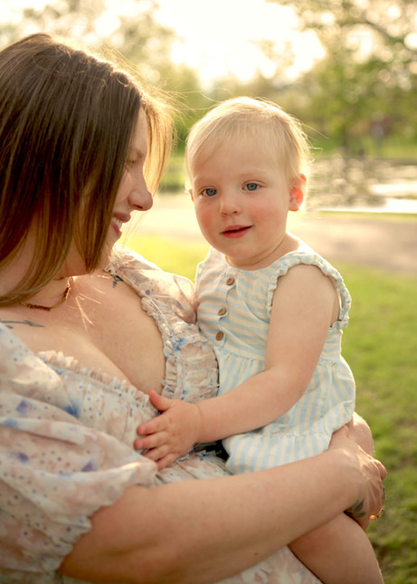 A pregnant mother is holding her toddler daughter and smiling, while backlit by golden hour light in Allegheny Commons Park.