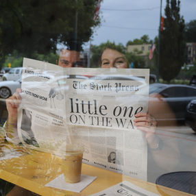 TR + Ava excitedly peek over a newspaper called "The Stork Press" and the headline "Little One On The Way" over coffee and through the window.