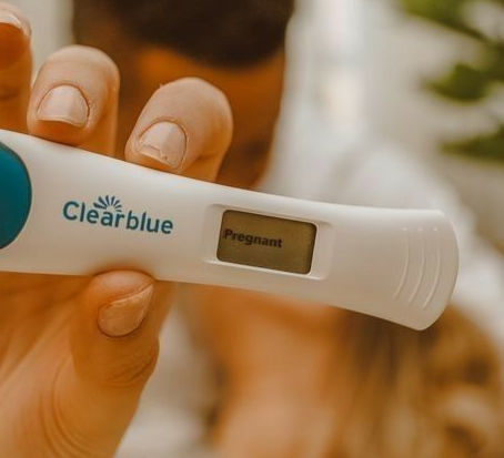 A close-up of a hand holding a Clearblue pregnancy test showing "Pregnant." Blurred background with greenery, warm tones, and a joyful mood.