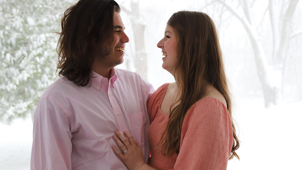 Ava and TR, both in pink, smile at each other, standing on a porch against a snowy backdrop, conveying warmth and happiness.