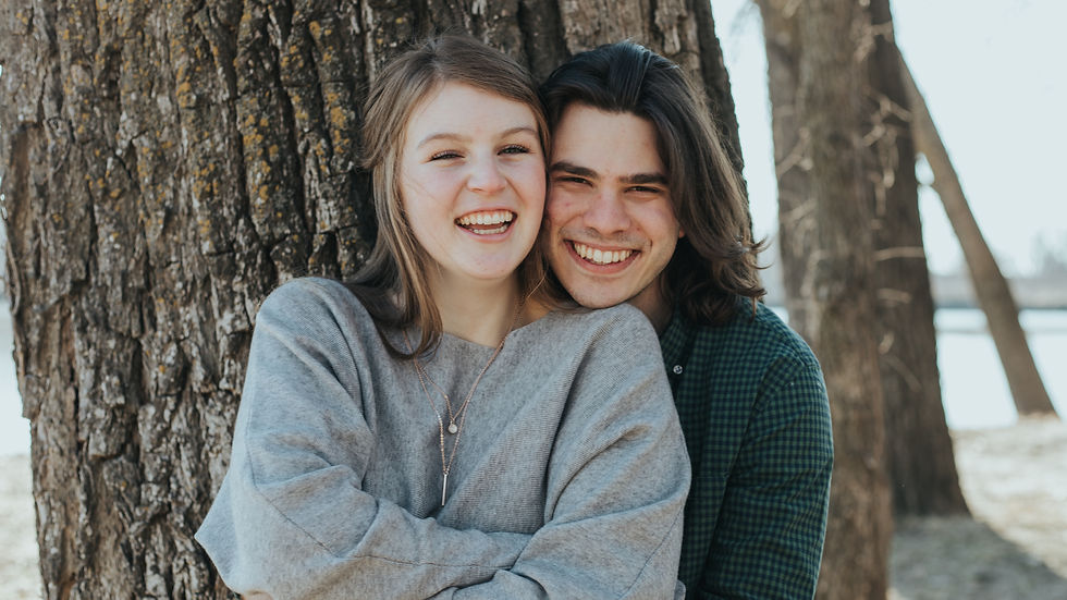 A smiling Ava and TR embrace in front of a tree, with Ava in a gray sweater andTR in a green plaid shirt, outdoors.