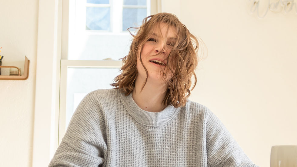 Ava smiles with tousled hair in a cozy gray sweater, seated indoors beside a sunlit window, exuding a joyful and playful mood.