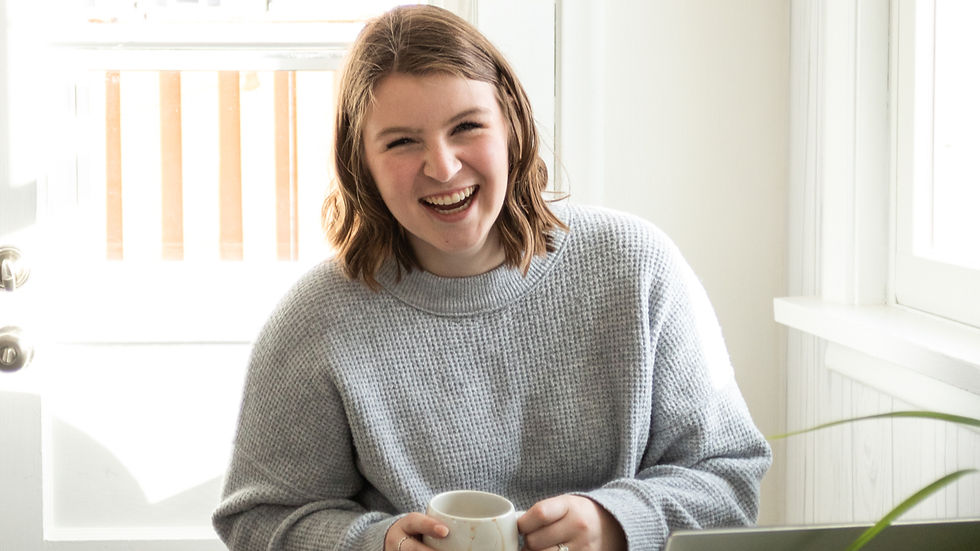 Ava in a gray sweater laughing boldly, holding a white mug. Sitting by a window with bright light, creating a cheerful, cozy atmosphere that screams of freedom.