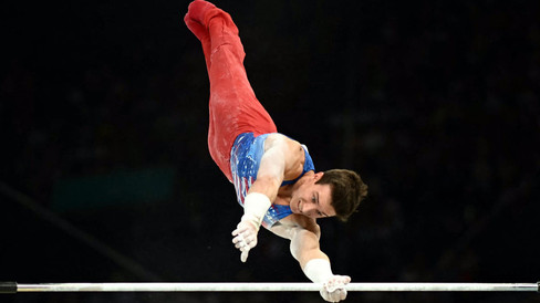 Gymnast Brody Malone swings around the high bar with one hand while the other hand reaches to make contact with the bar