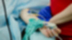 Female patient's hand with pulse oximeter, IV line, and tourniquet. Blue and red medical attire in background, suggesting a clinical setting.
