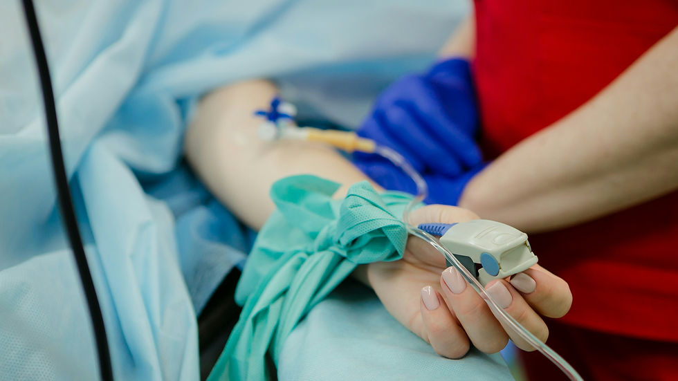 Female patient's hand with pulse oximeter, IV line, and tourniquet. Blue and red medical attire in background, suggesting a clinical setting.