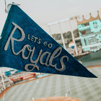 Hand holding a blue pennant reading "Let's Go Royals" above a baseball field. Stadium seats and scoreboard visible in the background.