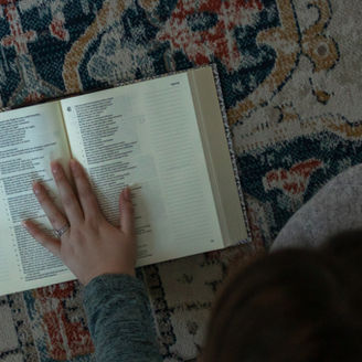Ava's left hand on an open Bible resting on a colorful carpet with blue, yellow, and red patterns. Pensive, serious, and focused setting.