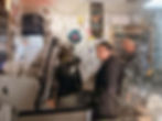 Three people in a cozy radio studio filled with posters and notes. One person smiles at the camera. Computers and recording equipment visible.