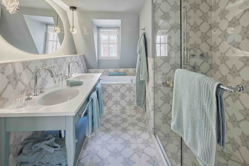 Badezimmer der Majesty Suite mit eleganten Fliesen, Doppelwaschbecken und Badewanne unter einem kleinen Dachfenster.