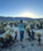 Woman holding a GoPro on a small tripod behind her back, walking through a field of cholla cacti.