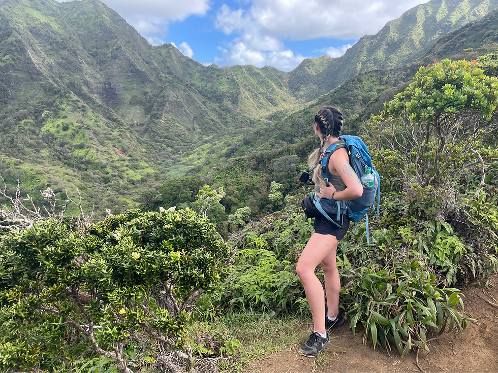 Woman with backpack on a trail looking toward lush, green mountains.