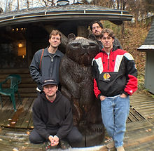 Four men, one crouching, on a wooden deck, rustic house backdrop.