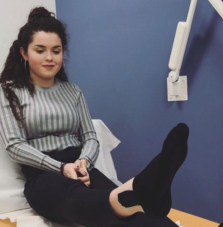 Niamh is sat on a bed in a doctors office, raising one leg.