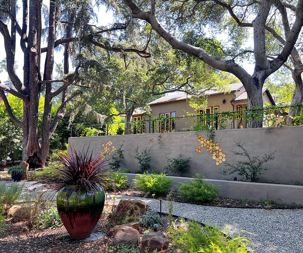 Gravel path beneath mature trees beside a stucco garden wall with mosaic tile accents and a glazed ceramic pot