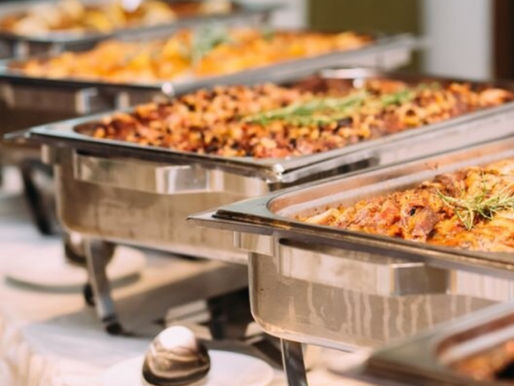 Row of stainless steel catering chafing dishes with warm dishes, including seasoned meats and roasted potatoes, displayed on a buffet table.