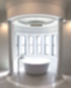 Elegant bathroom with a round white tub centered between columns, under a circular chandelier. Large windows offer natural light. Luxurious feel.