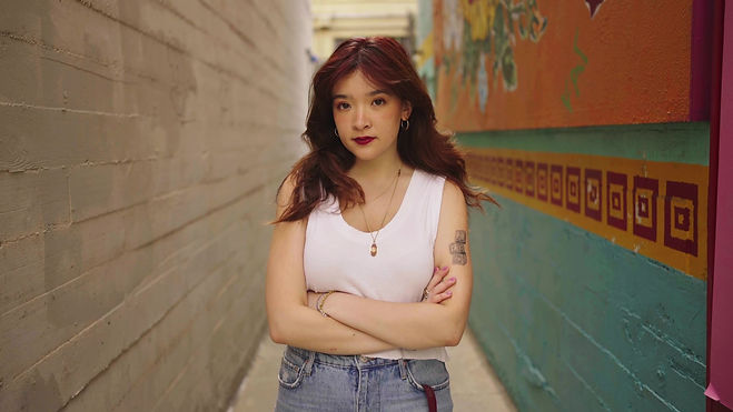 Photo of AML. She has long shoulder-length wavy red hair, wears a white tank top, jeans. She looks seriously at the camera.