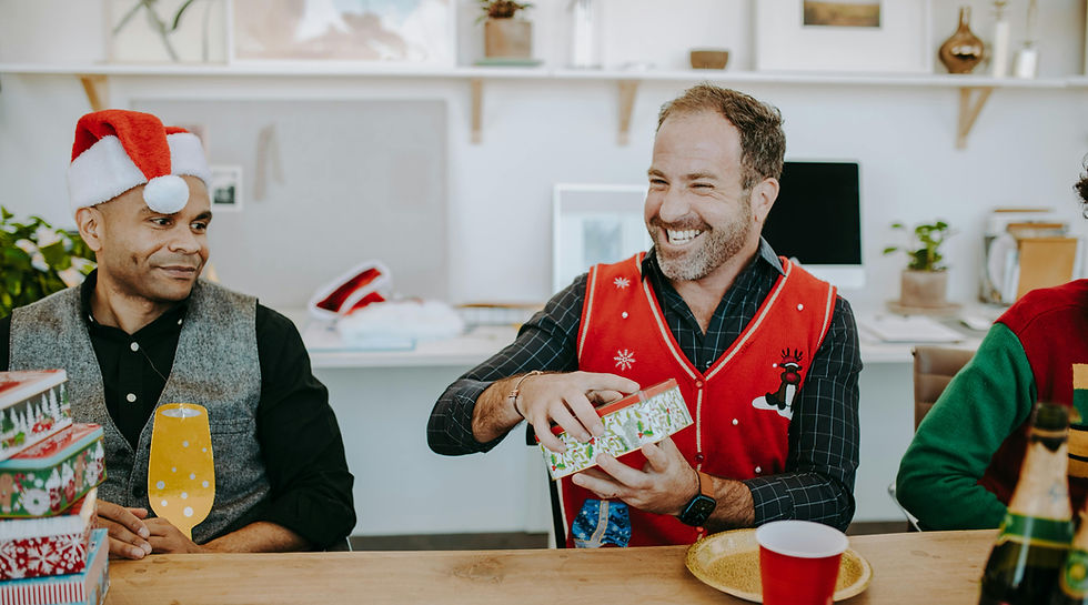 “Man in professional office attire wearing festive Christmas accessories, laughing while opening presents in a modern office setting
