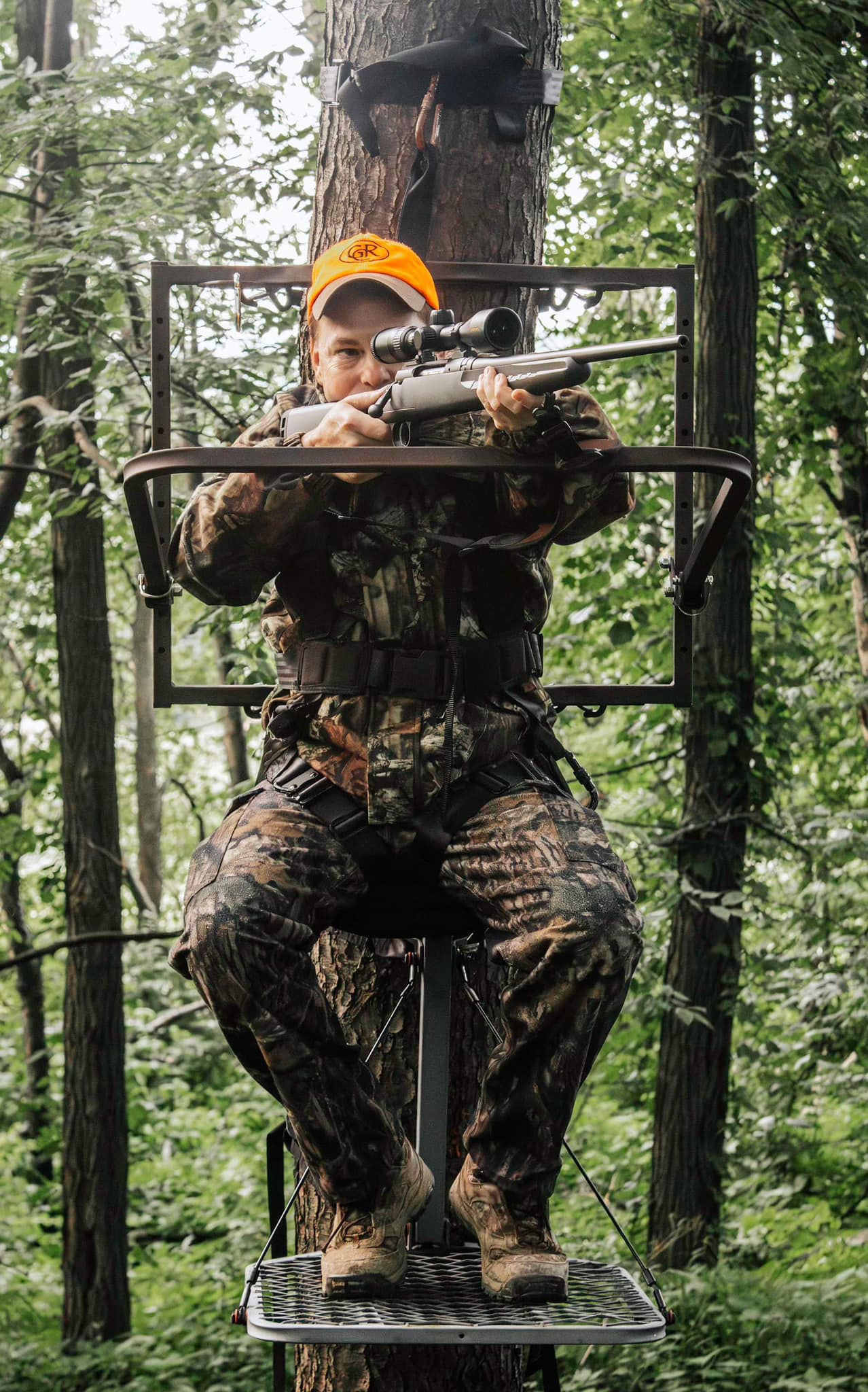 Hunter in camouflage, resting gun on treestand shooting rail. Looking down gun scope.