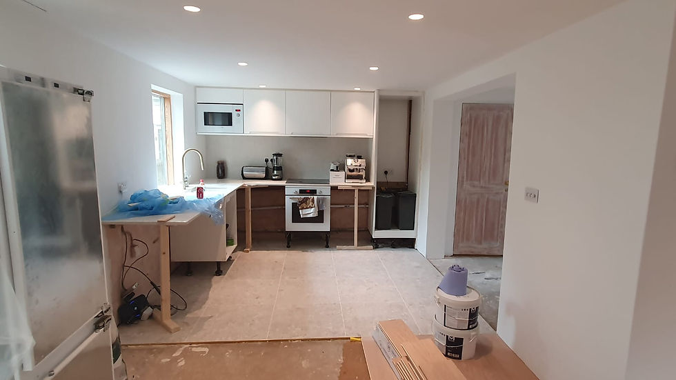 Renovated kitchen with white cabinets, unfinished lower units, and appliances. Paint cans and a wooden door in the background. Bright lighting.