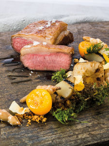 Rosa gebratenes Fleisch mit knuspriger Kruste, angerichtet mit buntem Gemüse, Kräutern und kleinen gelben Tomaten auf dunkler Holzplatte im Fine-Dining-Stil.