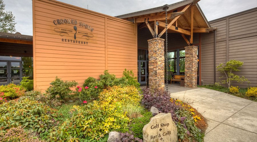 Entrance to Crooked Spoon restaurant with wooden siding and stone pillars. Bright flowers and shrubs in the foreground. Sign reads "Golf Shop Bar & Grill."