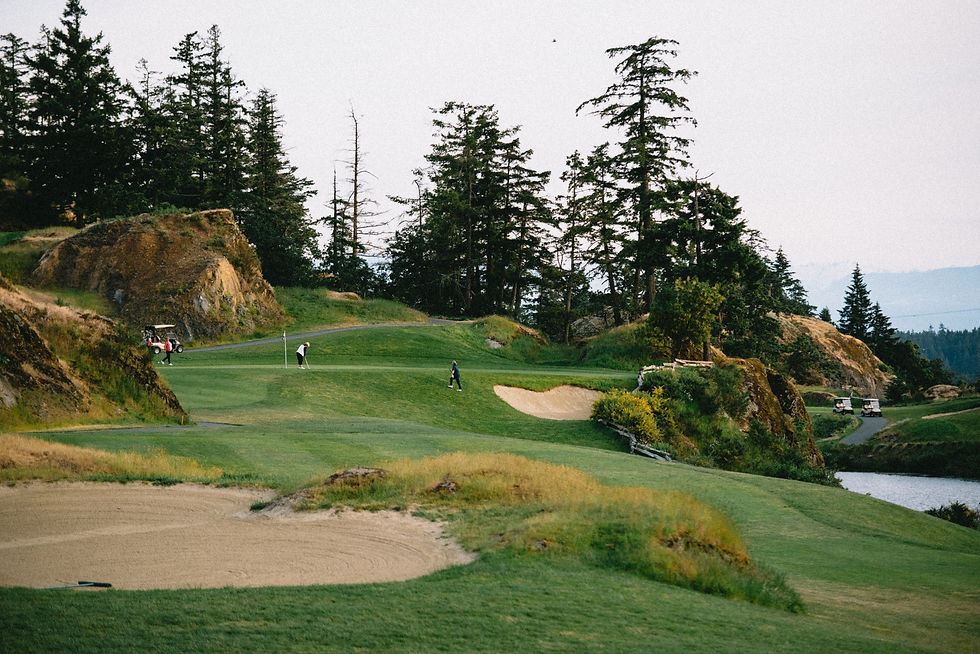 Golfers on a lush green golf course with rocky hills and trees in the background. Sand trap and golf carts visible. Peaceful, scenic setting.
