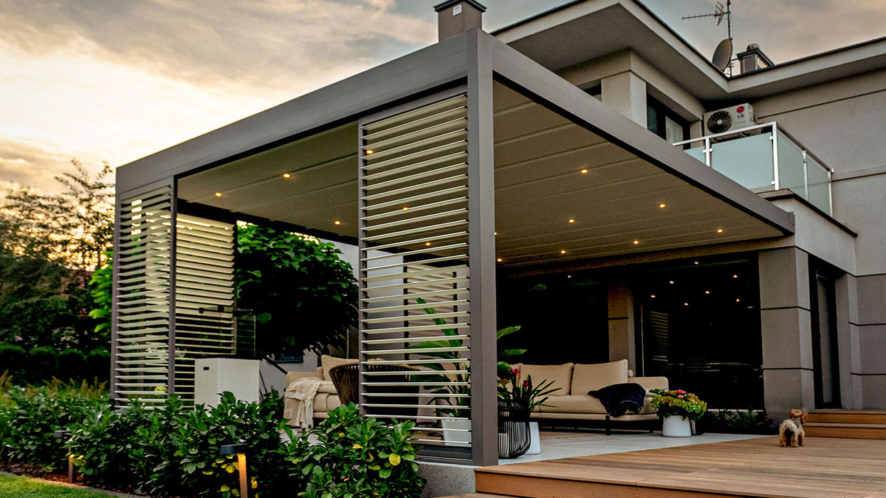Moderne Screenroom Lamellendach-Pergola mit seitlichen Design-Lamellen und integrierter LED-Beleuchtung auf einer großzügigen Holzterrasse. Die anthrazitfarbene Aluminiumkonstruktion erweitert den Wohnbereich eines modernen Einfamilienhauses und schafft einen stilvollen, wettergeschützten Outdoor-Loungebereich.