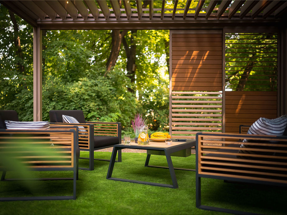 Gemütlicher Outdoor-Loungebereich unter einer Pergola mit festen Lamellen und seitlichen Holzlamellen. Moderne Gartenmöbel, natürliche Materialien und die grüne Umgebung schaffen einen geschützten, stilvollen Rückzugsort im Garten.