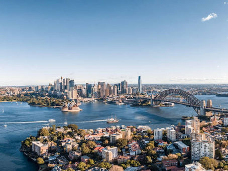 This image shows Sydney Harbour with the iconic Sydney Opera House and Harbour Bridge, surrounded by city skyscrapers, waterfront buildings, and boats sailing on a bright, clear day.
