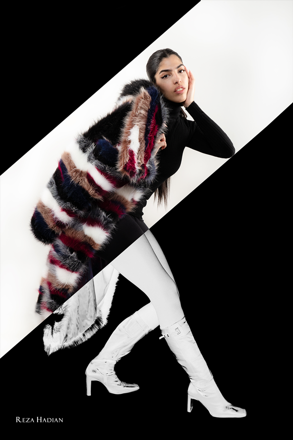 A fashion model is striking a pose in an artistic split-image composition. The upper part of the image features the model in a vibrant, multicoloured fur coat designed by Alisa Vaseghia over a black top, set against a white background. The lower part of the image shows the model in monochrome, wearing white thigh-high boots, with the background inverted to black. The split diagonal line creates a dynamic contrast between the colourful upper section and the stark black-and-white lower section.