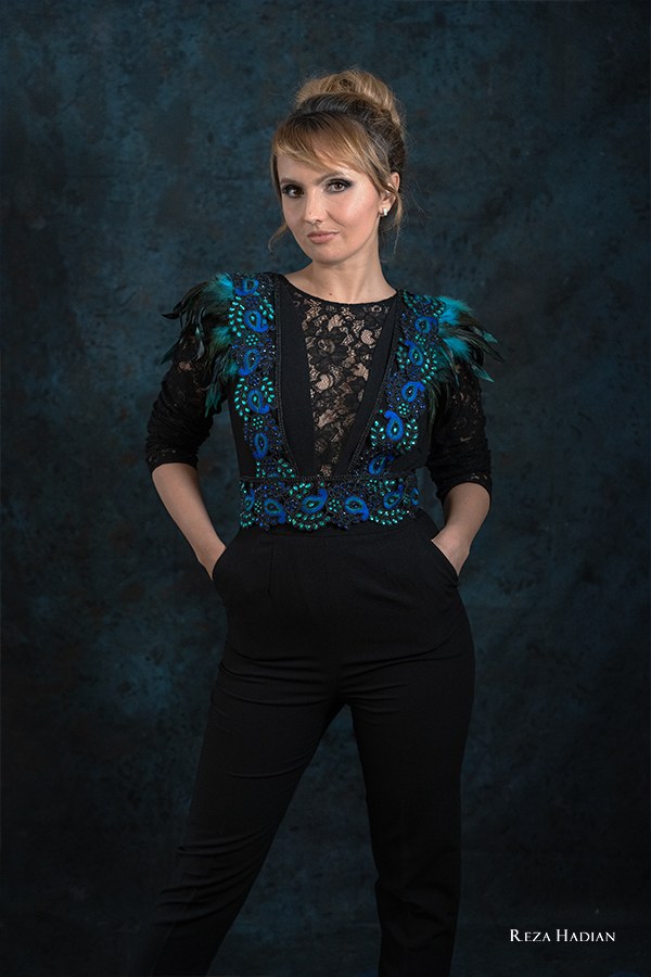 A confident woman poses against a dark, textured backdrop, wearing a stylish black lace top adorned with blue and green feathered accents and intricate beadwork. Her hair is elegantly styled in a bun, and she has a poised, self-assured expression with her hands resting in the pockets of her black pants.