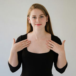 A woman stands against a plain light background, looking at the camera with a slight smile. She is wearing a black top and has both hands raised toward her upper chest, palms facing inward, as if demonstrating a sign.