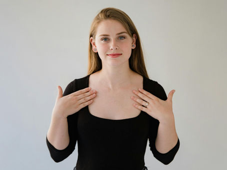 A woman stands against a plain light background, looking at the camera with a slight smile. She is wearing a black top and has both hands raised toward her upper chest, palms facing inward, as if demonstrating a sign.