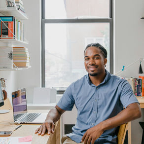 A man sits at a desk in a bright, modern office space. He is smiling and wearing a light blue short-sleeve button-down shirt and khaki pants. A laptop, papers, and books surround him, and a large window behind him shows an outside view.