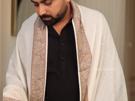 The image features a sophisticated man draped in an elegant Pashnoor Pashmina Shawl, exuding understated luxury and cultural refinement. The shawl is crafted in a pristine ivory-white hue with an intricate beige border featuring traditional Kashmiri motifs in fine weave. His all-black kurta creates a bold contrast, allowing the soft-toned shawl to shine with subtle grace. The man’s contemplative pose and refined styling add a modern charm to this classic ethnic attire. The Pashnoor logo in the top right corner and at the bottom reinforces the exclusivity of the brand.