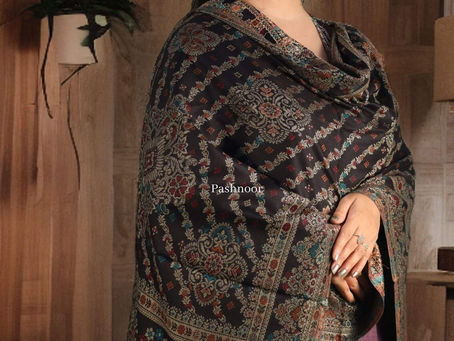 The image features a graceful woman draped in an intricately woven shawl from Pashnoor. The shawl displays a rich black base adorned with traditional Kashmiri-style motifs in shades of maroon, teal, and gold. Its elaborate floral and paisley patterns reflect timeless elegance and fine craftsmanship. The background features a cozy wooden interior with soft lighting, enhancing the luxurious feel of the shawl. The Pashnoor logo is placed on the top left corner, and the brand name "pashnoor" is written at the bottom in an elegant serif font.