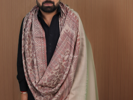 The image portrays a dignified gentleman wrapped in a sophisticated Pashnoor shawl. The shawl features a beautiful gradient of soft pink and cream shades, creating an elegant and serene look. The delicate weave and gentle colors evoke a sense of warmth, tradition, and understated luxury. Set against a muted brown background, the shawl stands out while the Pashnoor logo subtly reinforces the brand’s commitment to heritage and craftsmanship.