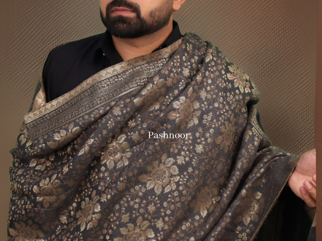 A confident man models an elegant, dark-toned Pashmina shawl from Pashnoor, draped regally across his shoulders. The shawl features a luxurious, intricate paisley and floral pattern in earthy hues—rich browns, muted golds, and subtle olive greens—showcasing the depth and heritage of Kashmiri craftsmanship. He stands against a warm, neutral background, enhancing the shawl’s texture and royal appeal. The Pashnoor logo in the top left corner adds a refined brand presence.